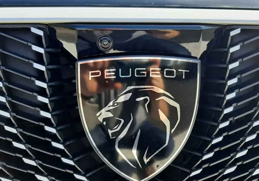 Gros plan sur la calandre noire et le logo Peugeot lion argenté du 3008 blanc Okenite, avec caméra intégrée au-dessus.