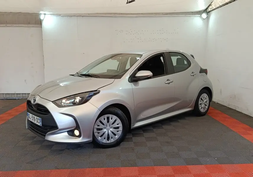 Toyota Yaris hybride gris clair vue 3/4 avant droit dans un garage avec feux allumés et jantes standard.