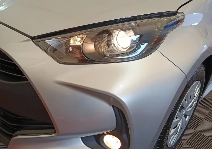 Gros plan sur l'avant droit gris clair de la Toyota Yaris hybride 2022, mettant en valeur les phares allumés et le feu antibrouillard.