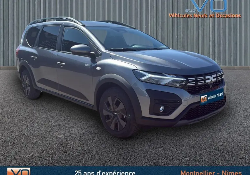 Vue 3/4 avant d'un Dacia Jogger gris 2025 avec barres de toit et jantes spécifiques, stationné sur sol goudronné.