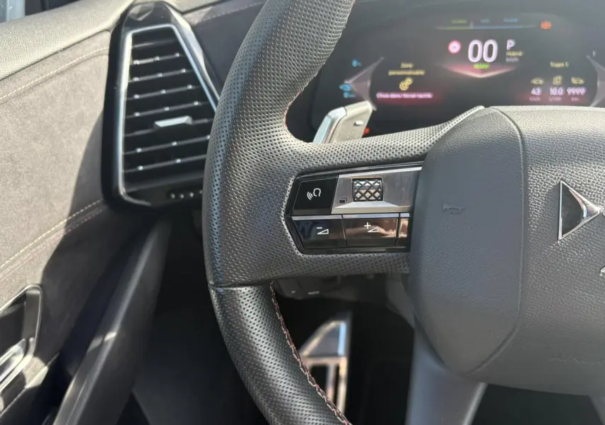 Gros plan sur le volant perforé noir avec surpiqûres Carmin et Or du DS7 1.6 E-Tense 2024, tableau de bord numérique en arrière-plan.