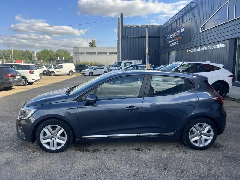 Profil droit d'une Renault Clio 1.6 E-Tech 140ch Business gris Titanium garée devant un bâtiment industriel.