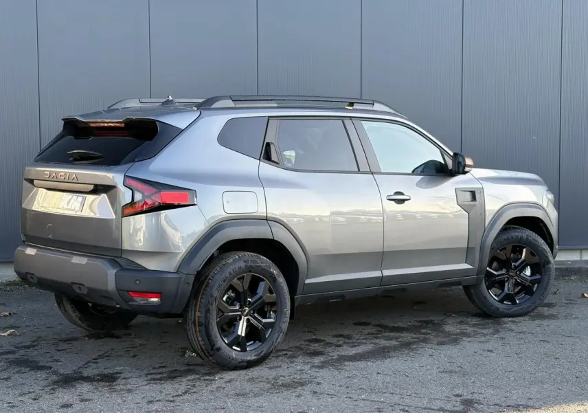 Vue 3/4 arrière droite du Dacia Duster 2026 gris schiste avec jantes alliage noires et barres de toit longitudinales.