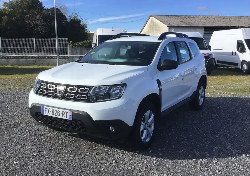 Dacia Duster blanc en 3/4 avant droit, avec calandre noire et jantes alliage 16 pouces, stationné sur un sol gravillonné.