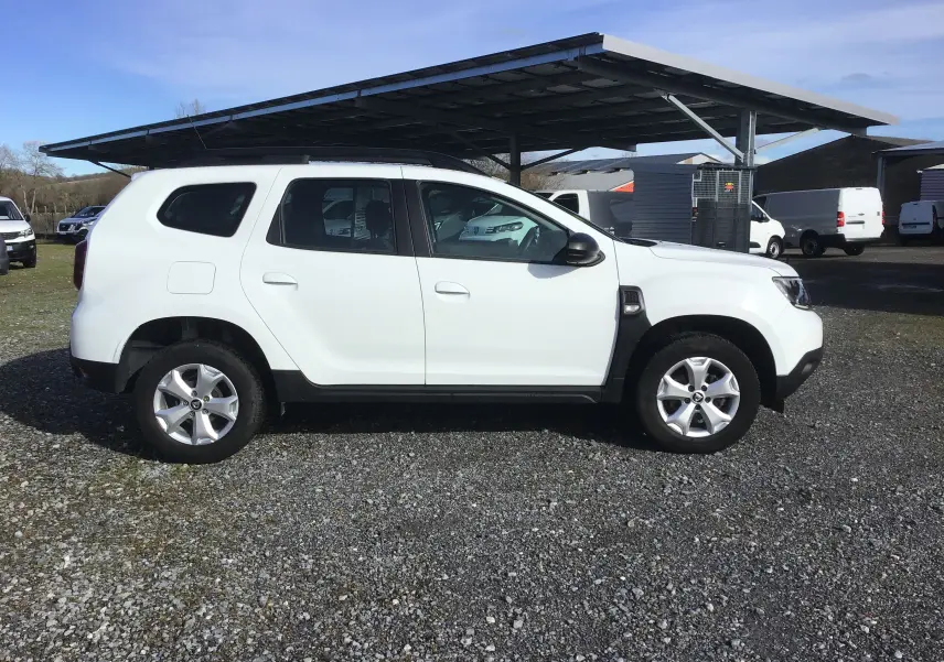 Profil côté gauche d'un Dacia Duster blanc 2021, version 4x2, avec jantes alliage et toit noir sous un abri extérieur.