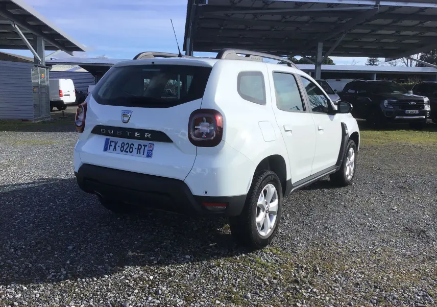 Vue 3/4 arrière droite d'un Dacia Duster blanc 2021 avec jantes alliage et barres de toit sous un abri.