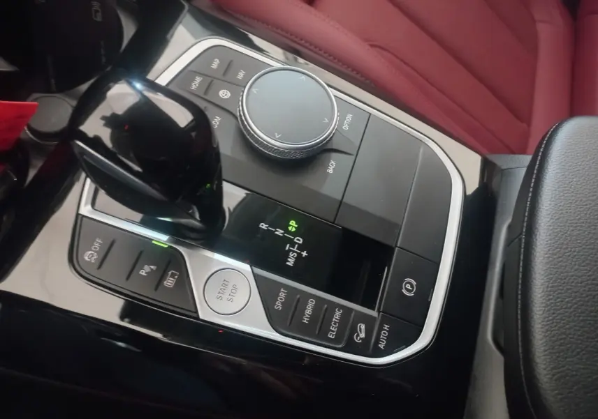 Vue rapprochée de la console centrale noire avec levier de vitesses et molette de sélection, sièges en cuir rouge visibles.