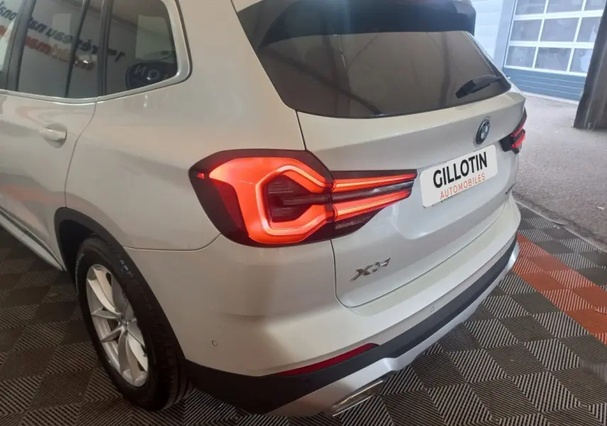 Vue 3/4 arrière gauche d’un BMW X3 blanc avec feux arrière allumés et logo visible sur coffre.