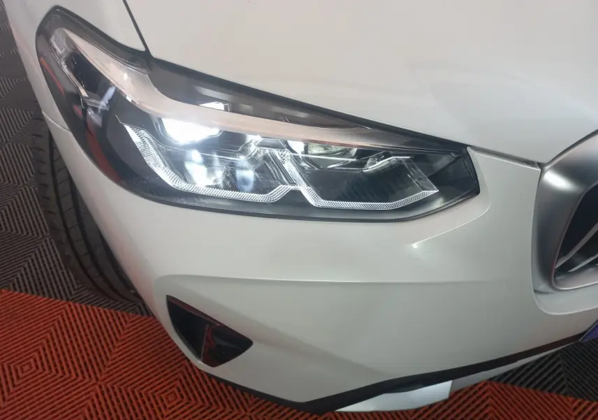 Gros plan sur le phare avant droit allumé d’un BMW X3 blanc, avec partie de la calandre visible.