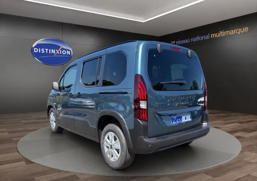 Vue 3/4 arrière droite d'un Peugeot Rifter bleu 2026 avec feux arrière LED et jantes argentées en showroom.