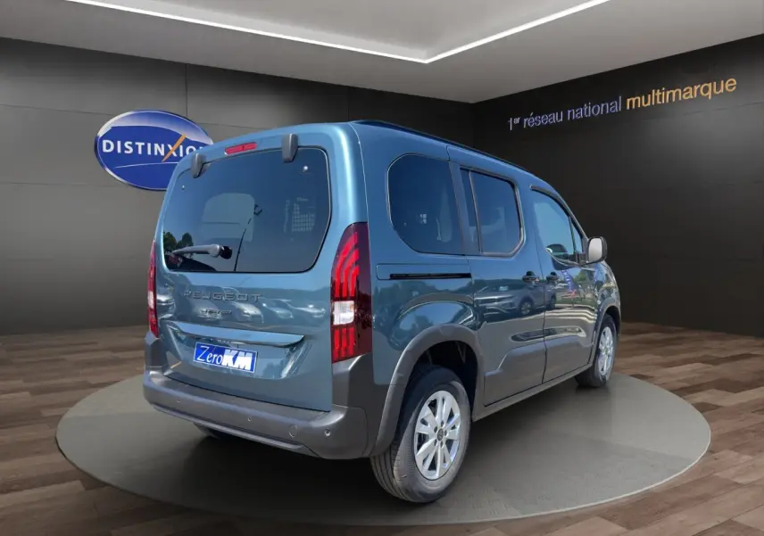 Vue 3/4 arrière droite du Peugeot Rifter bleu 2026 avec feux arrière verticaux et hayon vitré dans un showroom.
