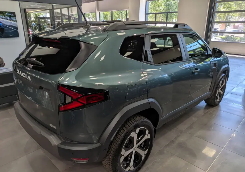 Vue 3/4 arrière droite d’un Dacia Duster Hybrid-G 150 CH 4x4 vert cèdre avec jantes biton et barres de toit en showroom.