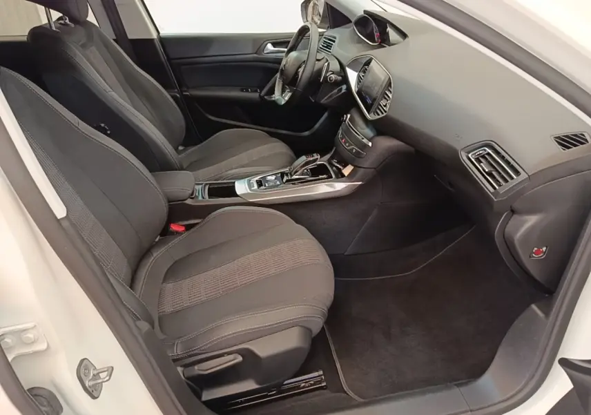 Intérieur avant droit de la Peugeot 308 blanche, montrant sièges noirs, tableau de bord avec écran tactile et volant cuir.
