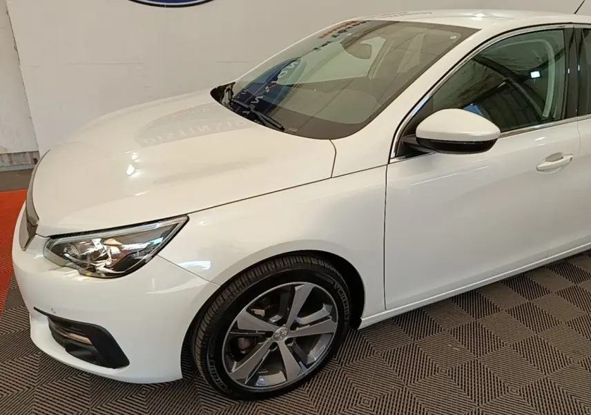 Vue 3/4 avant droit d'une Peugeot 308 blanche avec jantes alliage et feux avant LED visibles en intérieur showroom.