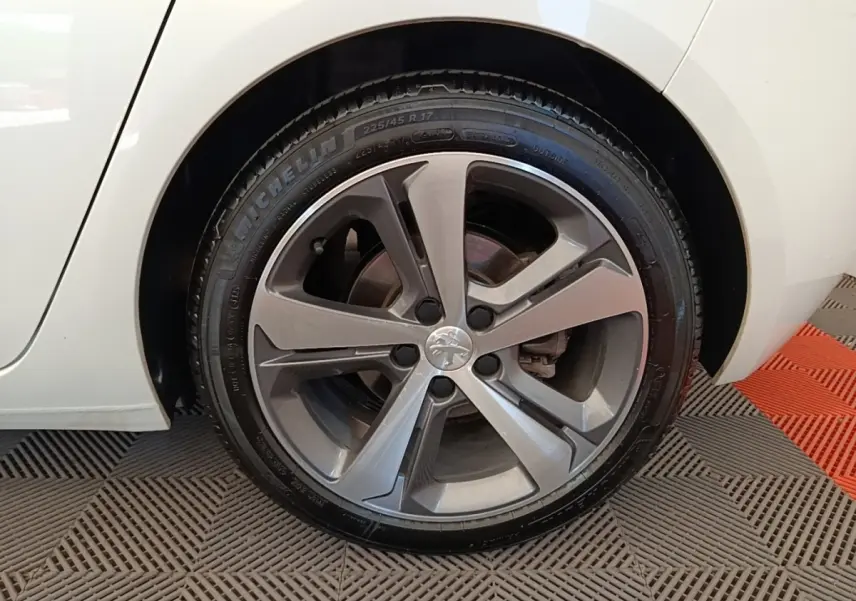 Gros plan sur la roue avant droite d'une Peugeot 308 blanche, jante alliage gris foncé au design moderne.
