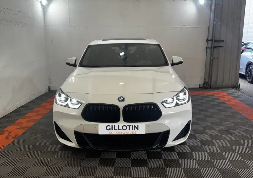 BMW X2 xDrive25e blanc vue de face avec phares LED allumés et calandre noire sportive dans un garage.