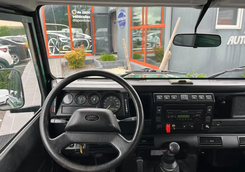 Vue intérieure frontale du tableau de bord noir du Land Rover Defender 90 vert, avec volant, compteurs et levier de vitesses manuel.