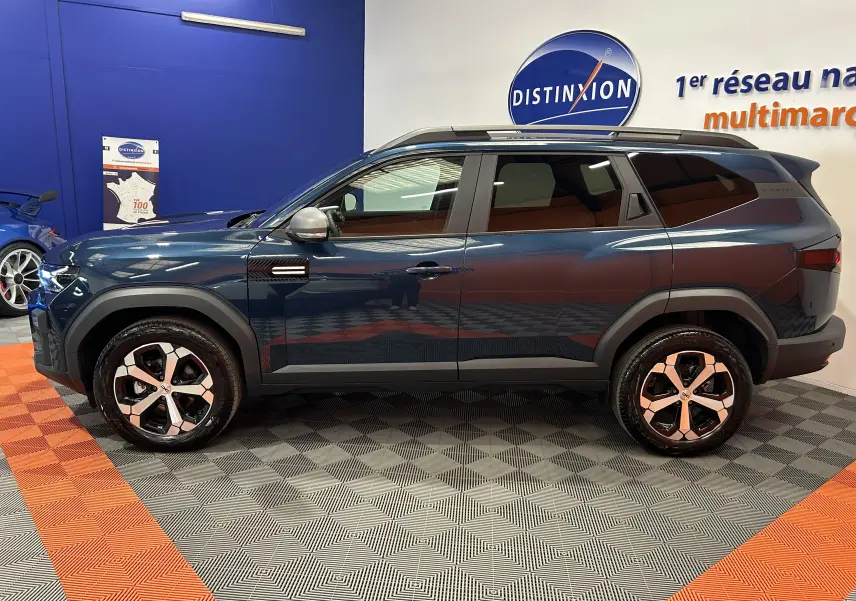 Vue de profil côté gauche du Dacia Bigster bleu indigo avec jantes bi-ton et barres de toit noires en intérieur showroom.