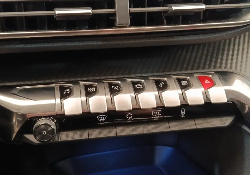 Gros plan sur la rangée de boutons métalliques du tableau de bord du Peugeot 5008 noir, avec bouton rouge d’alerte.