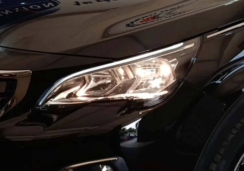Gros plan sur le phare avant droit allumé d’un Peugeot 5008 noir, avec reflet sur la carrosserie brillante.
