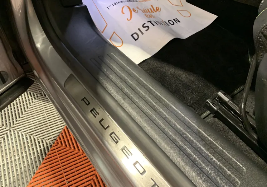 Gros plan sur la plaque de seuil de porte côté conducteur d'un Peugeot 2008 gris, avec tapis de sol et affiche partielle visible.