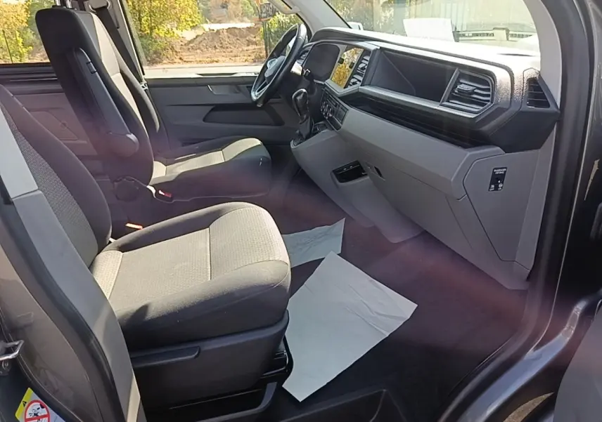 Vue intérieure côté conducteur du Volkswagen Transporter gris foncé, montrant sièges tissu et tableau de bord moderne.