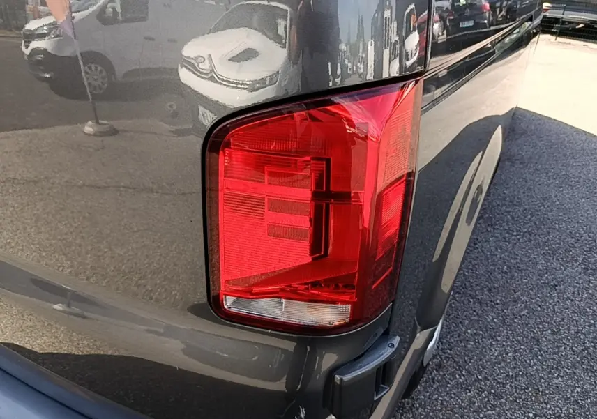 Gros plan sur le feu arrière droit du Volkswagen Transporter gris foncé, avec reflet de l’environnement urbain.