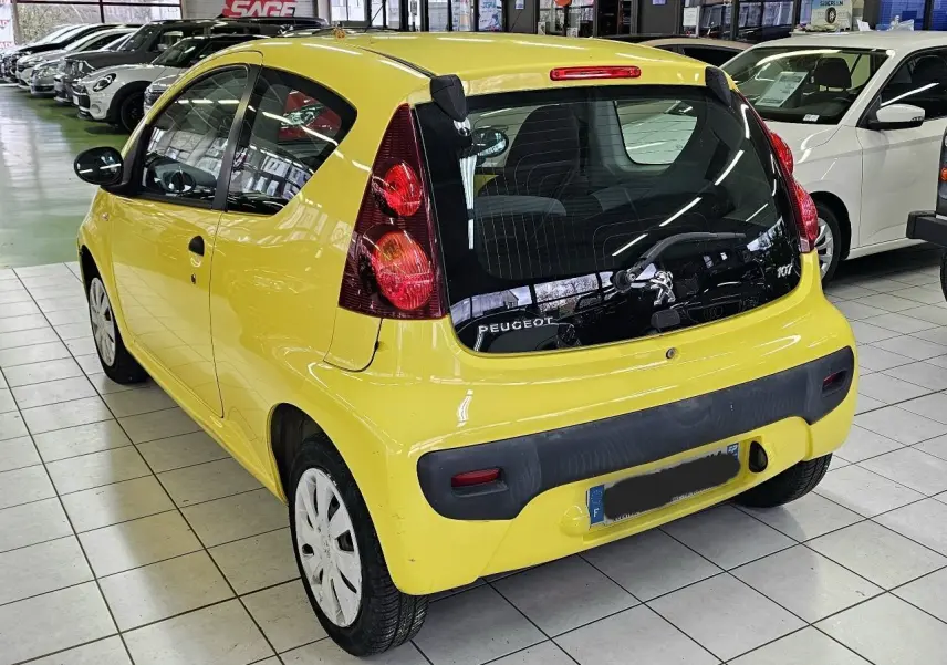 Vue 3/4 arrière droite d'une Peugeot 107 jaune vif en intérieur, avec feux arrière verticaux et pare-chocs noir.