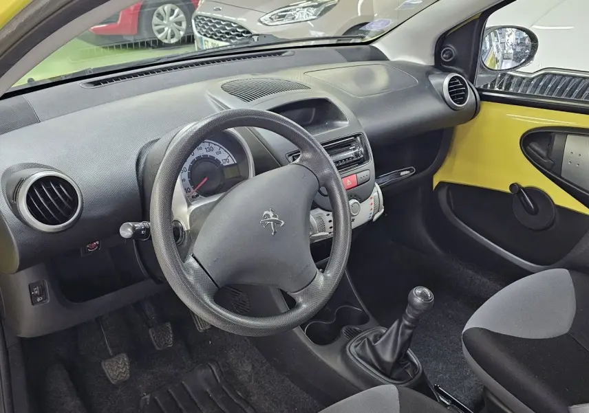 Intérieur côté conducteur d'une Peugeot 107 jaune de 2013, volant, tableau de bord et levier de vitesses manuelle visibles.