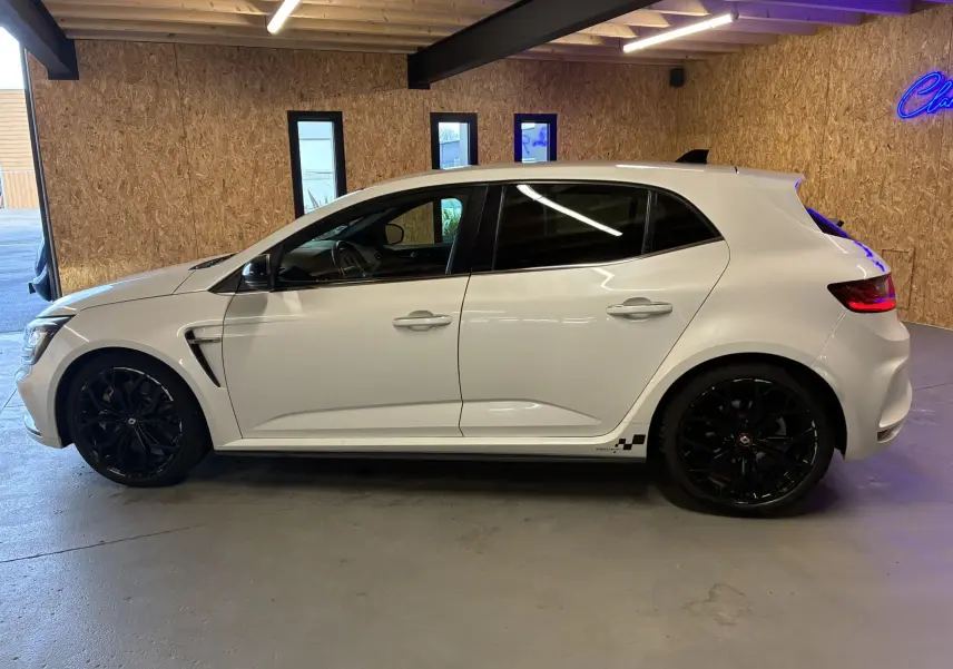 Profil côté gauche d'une Renault Mégane IV RS 280 EDC6 blanche avec jantes noires et détails sportifs au bas de caisse.