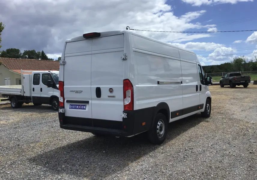 Fiat Ducato Fourgon blanc vu en 3/4 arrière droit sur un terrain gravillonné sous ciel partiellement nuageux.