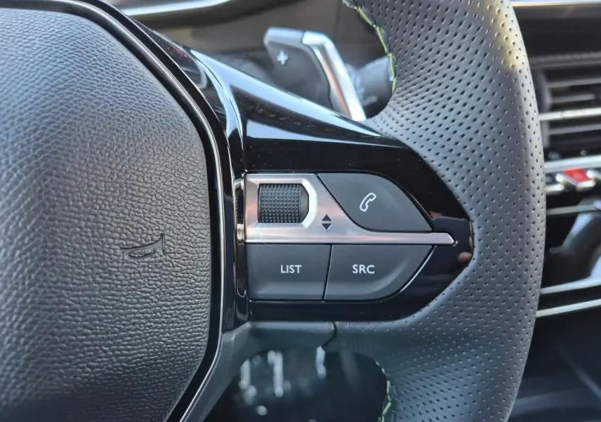 Gros plan sur le côté gauche du volant perforé noir de la Peugeot 208 Hybrid 136 e-DCS6 GT avec commandes audio intégrées.