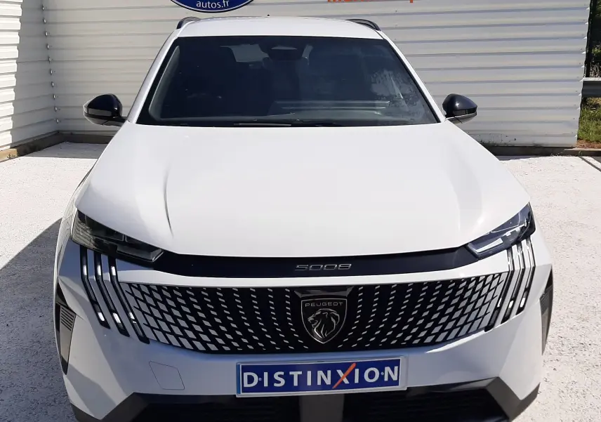 Vue avant d'un Peugeot 5008 blanc Okenite avec calandre noire distinctive et logo lion central, stationné devant un mur publicitaire.