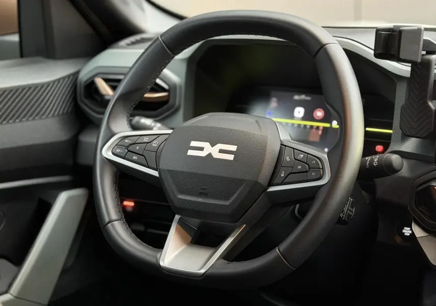 Vue rapprochée du volant noir du Dacia Duster 2025 avec commandes intégrées et tableau de bord digital en arrière-plan.