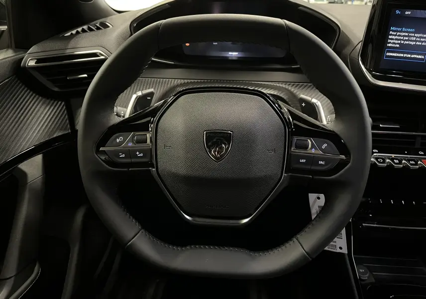 Vue rapprochée du volant noir au design hexagonal du Peugeot 2008 1.2 Hybrid 145ch, avec tableau de bord et écran tactile visibles.