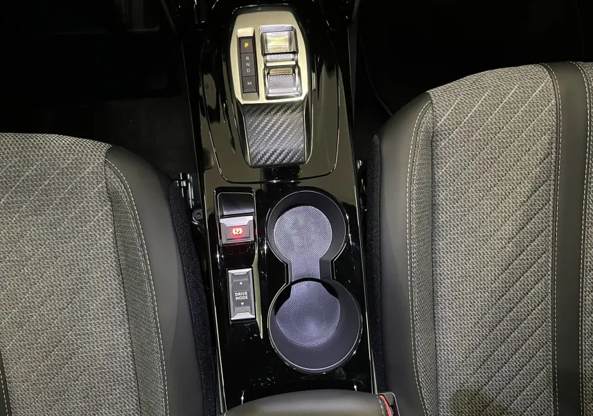 Vue plongeante sur la console centrale noire brillante du Peugeot 2008 2025, avec sélecteur de vitesse et porte-gobelets entre sièges gris tissu.