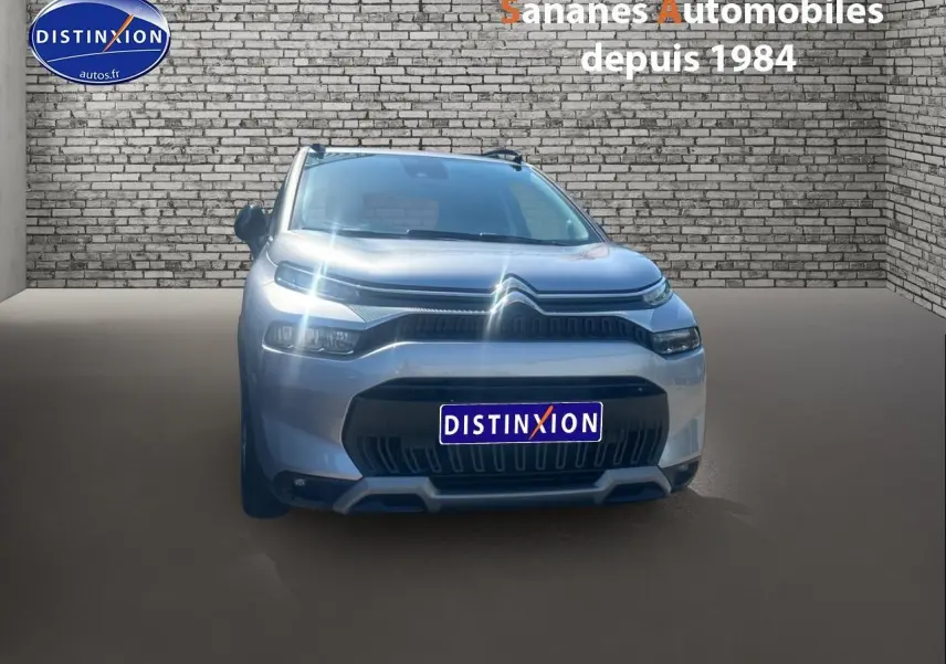 Vue frontale d'un Citroën C3 Aircross gris foncé avec projecteurs allumés, sur fond de mur en briques.