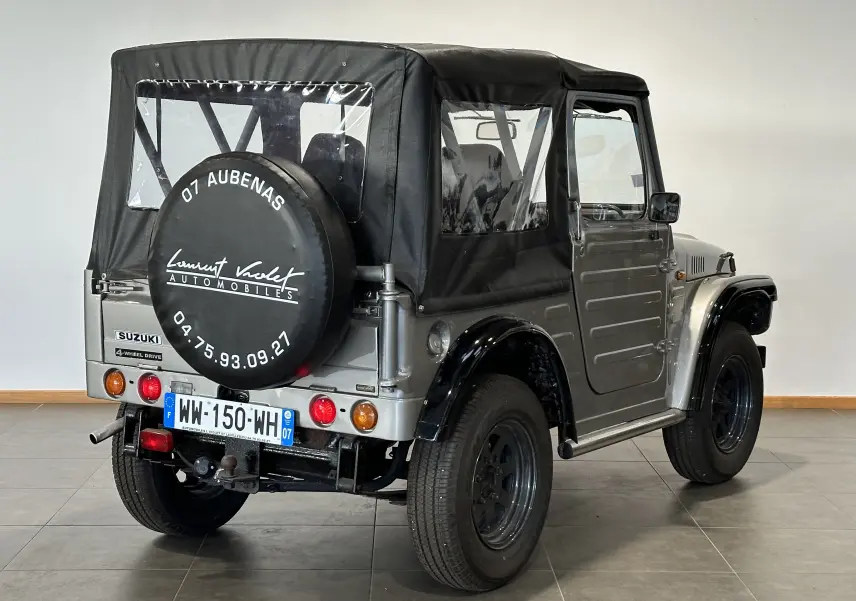 Vue 3/4 arrière droite d'un Suzuki Across LJ80 Cabriolet gris 1981 avec capote noire et roue de secours à l'arrière.