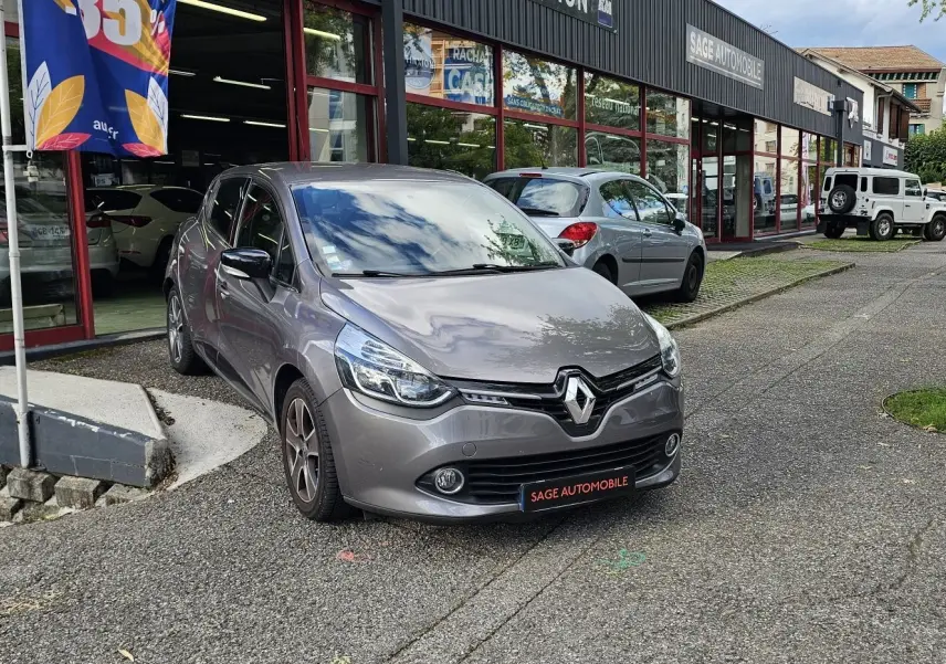 Renault Clio IV grise vue 3/4 avant droit, garée devant un garage avec plaque SAGE AUTOMOBILE.