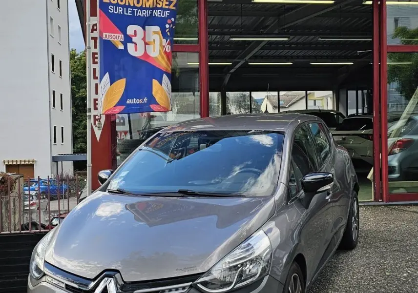 Renault Clio IV grise vue 3/4 avant droit devant un garage AD Expert avec drapeau promotionnel bleu.