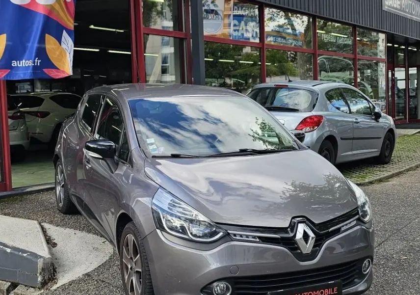 Renault Clio IV grise vue 3/4 avant droit devant un garage avec enseignes et panneau publicitaire coloré.