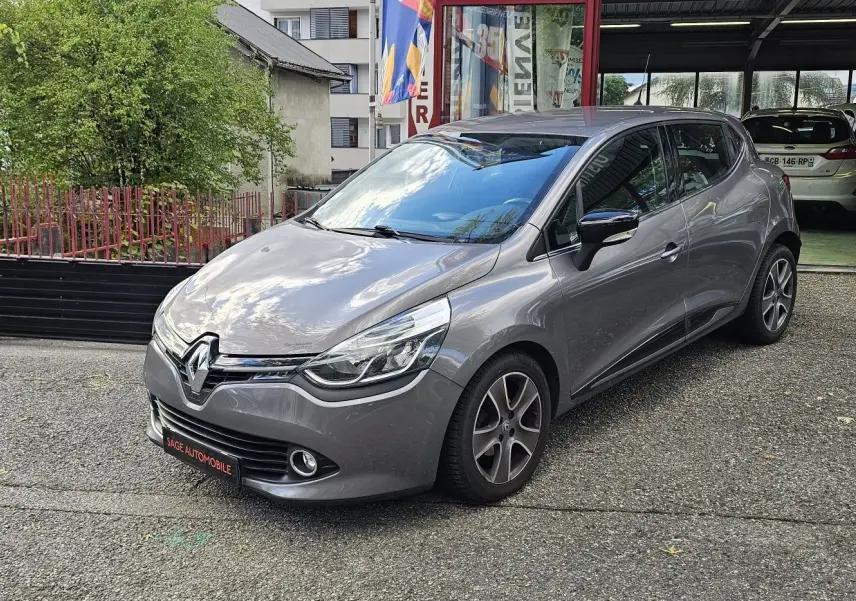 Renault Clio IV grise vue 3/4 avant droit, garée devant un bâtiment avec reflet du ciel sur le capot.