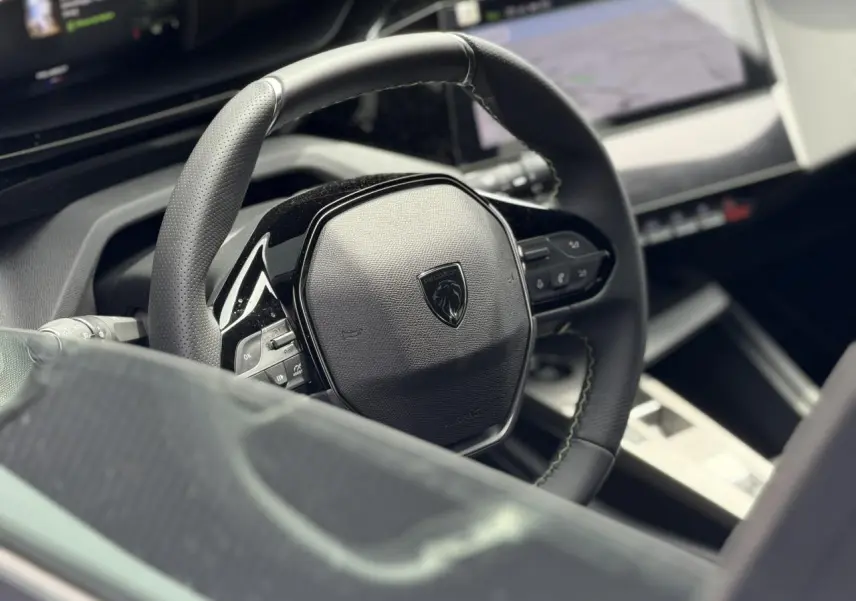 Vue rapprochée du volant compact noir du Peugeot 308 NV Hybrid 145 GT avec surpiqûres vert Adamite et tableau de bord numérique.