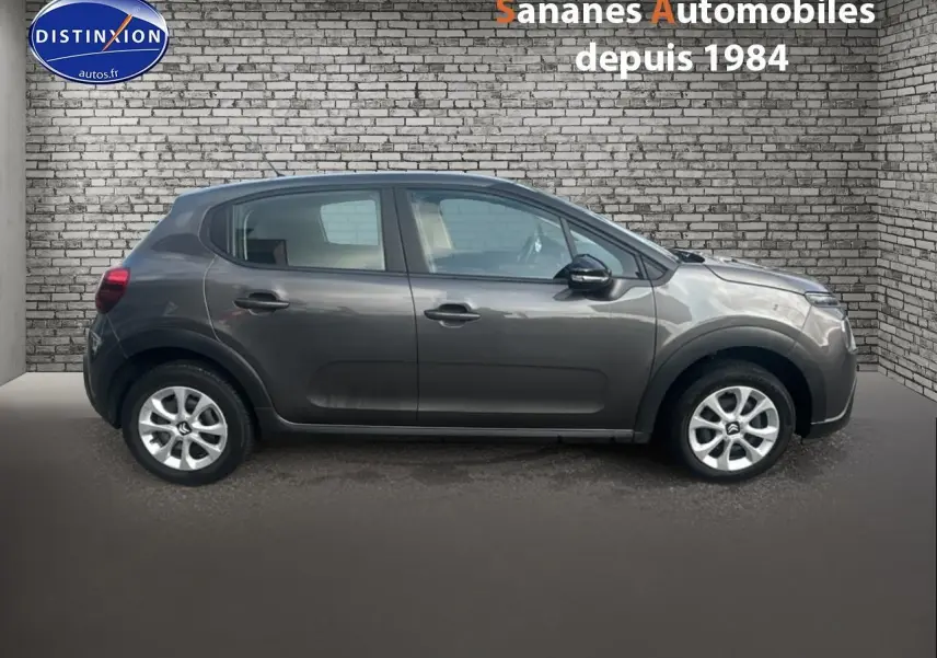 Profil côté gauche d'une Citroën C3 gris foncé 2023 avec jantes argentées, stationnée devant un mur en briques.