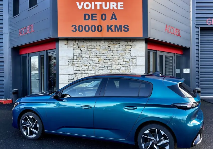 Peugeot 308 Hybrid 136 e-DCS6 Allure bleu vue de profil côté gauche devant un bâtiment avec panneau orange "Votre nouvelle voiture".