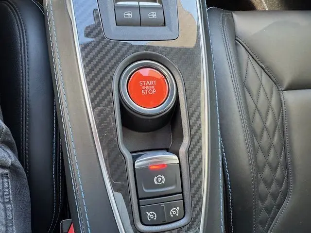 Gros plan sur la console centrale carbone de l'Alpine A110 gris acier avec bouton start/stop rouge et cuir noir surpiqué bleu.