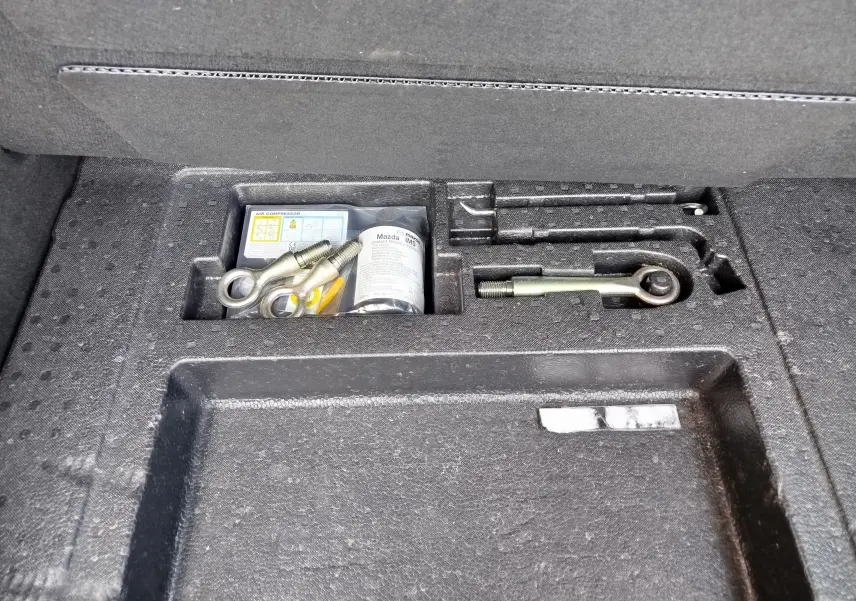 Gros plan sur le compartiment à outils du coffre du Mazda CX-30 noir, montrant les crochets de remorquage et accessoires rangés.