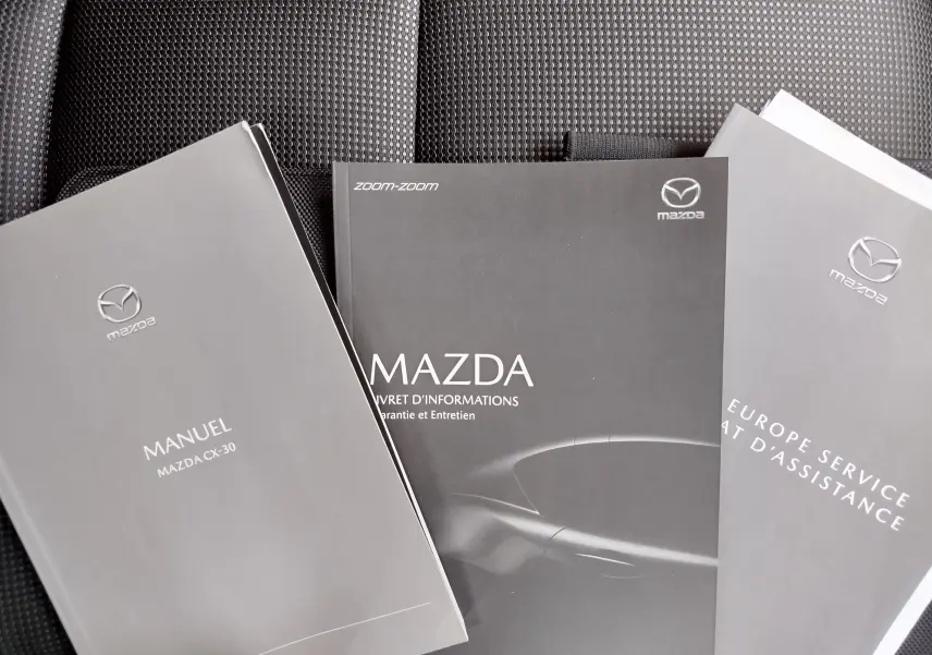 Trois manuels Mazda gris posés sur un siège en tissu noir, dont un manuel du CX-30 visible au premier plan.