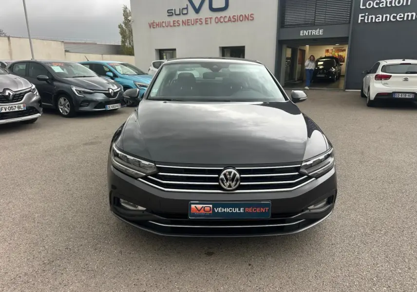 Volkswagen Passat gris foncé vue de face, avec calandre chromée et phares LED allumés sur parking extérieur.