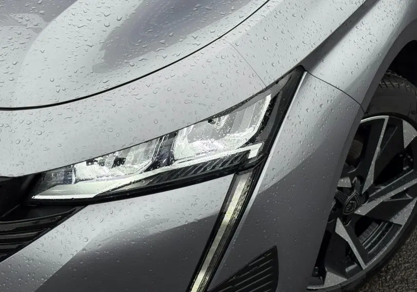 Gros plan sur le phare avant gauche et la roue alliage 17'' d'une Peugeot 308 SW Hybrid gris Artense sous la pluie.